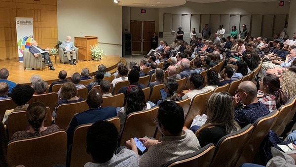 Crowd of attendees in an auditorium listens to a discussion with two men on a stage (Joseph Stiglitz and Ray Offenheiser).