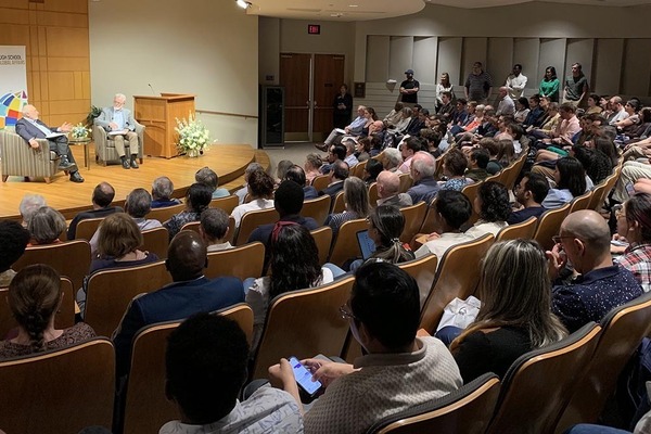 Crowd of attendees in an auditorium listens to a discussion with two men on a stage (Joseph Stiglitz and Ray Offenheiser).