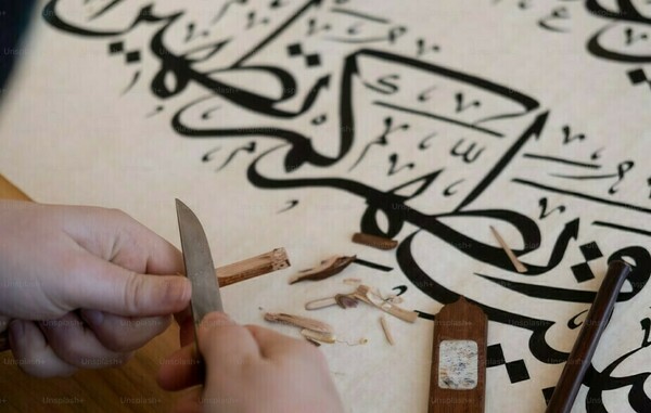 A person's hands, sharpening a wooden tool, with a sheet of Arabic Calligraphy.