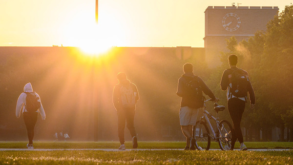 A group of students walk in the direction of an an academic building silhouetted by the sun.