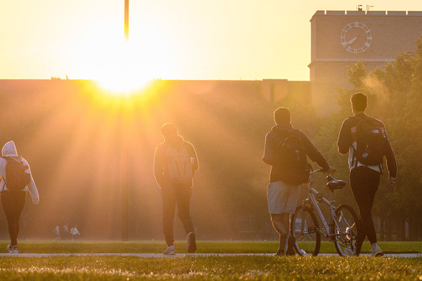 A group of students walk in the direction of an an academic building silhouetted by the sun.